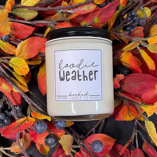 Candle labeled 'hoodie Weather' surrounded by autumn leaves and berries