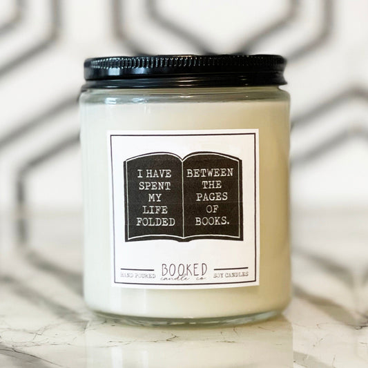 Image of a handmade candle in a glass container with a book label saying "I Have Spent My Life Folded Between The Pages Of Books"
