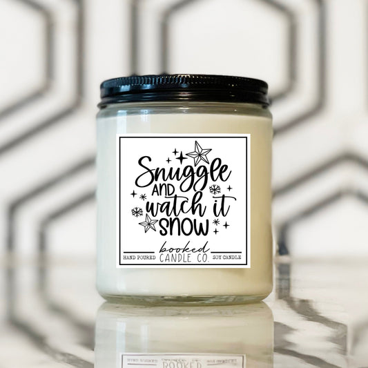Candle jar with a label reading 'Snuggle and watch it snow' on a reflective surface with a geometric background.