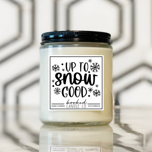 Candle jar with 'Up to Snow Good' label on a reflective surface with geometric background