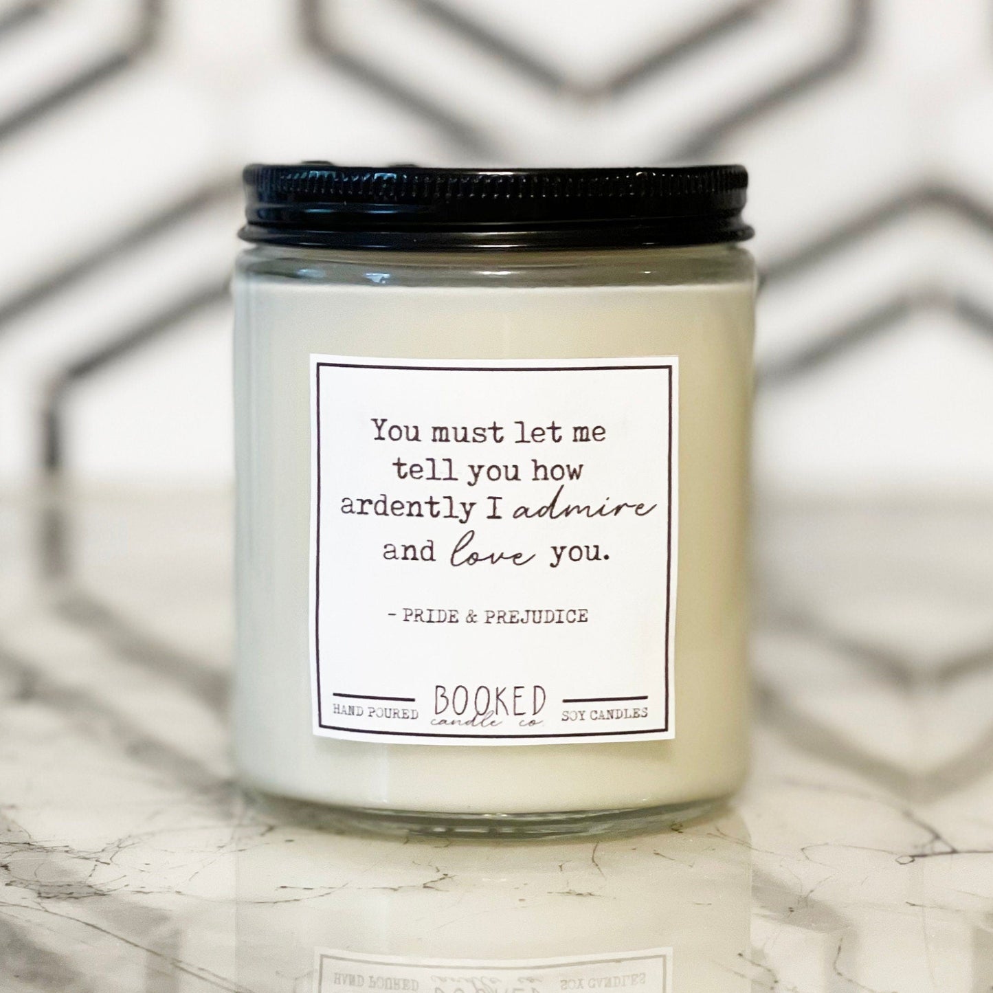 A white soy wax candle in an 8oz glass jar with a black lid, labeled 'Ardently Admire And Love - Pride & Prejudice' on a marble surface.