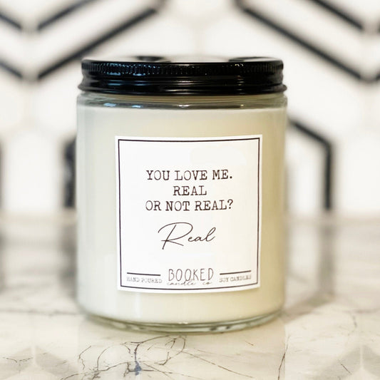 A white candle with a black lid, labeled 'You Love Me, Real Or Not Real - Hunger Games Series' placed on a table with a geometric patterned tablecloth.