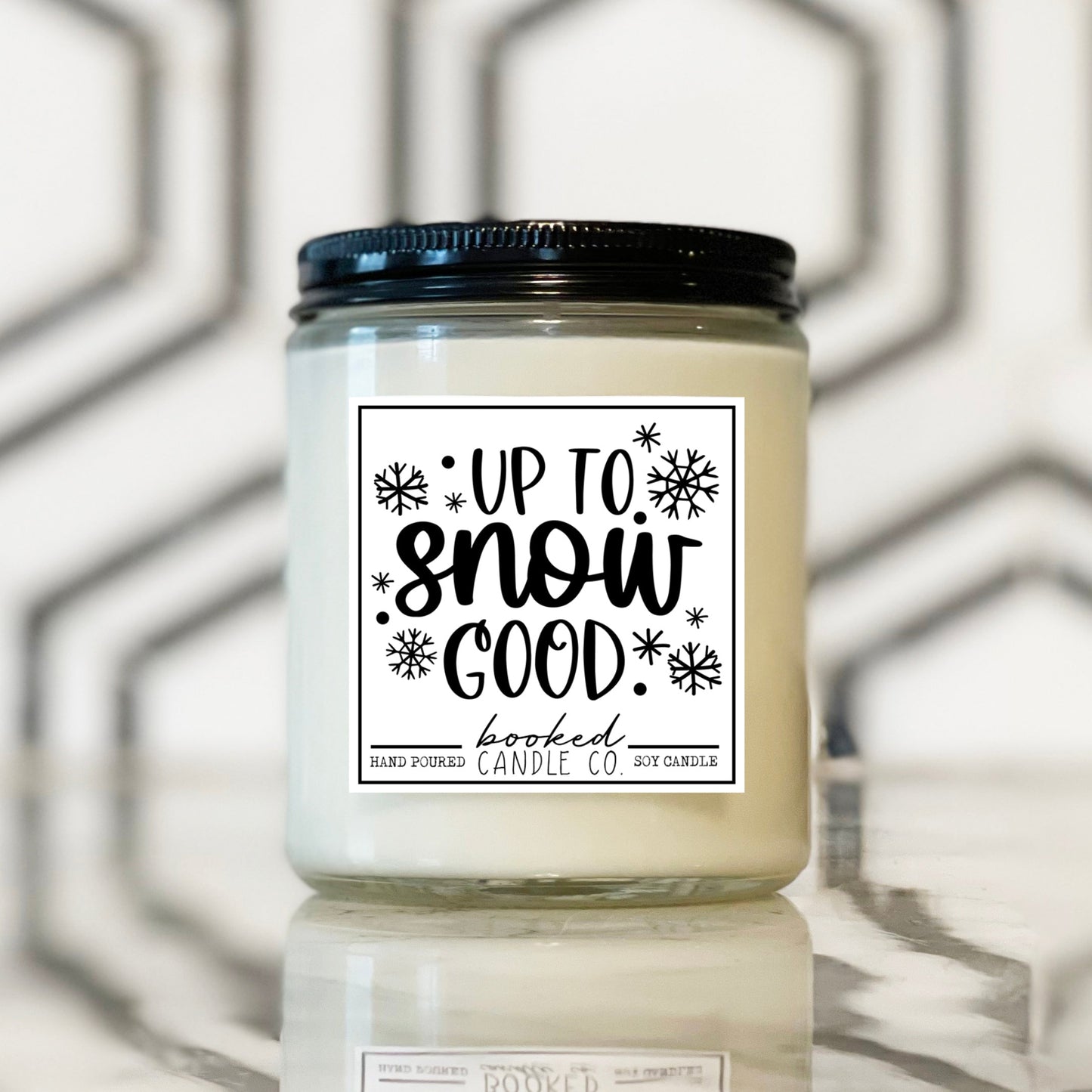 Candle jar with 'Up to Snow Good' label on a reflective surface with geometric background